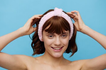 a cute woman with smooth radiant skin and dark silky beautiful hair stands on a blue background in a white towel with a pink bandage on her head after a bath. Horizontal studio photography.