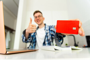 A man in a plaid shirt and glasses smiles and holds a credit card in his hands. A bank card in the hands of a young businessman. Focus on credit card