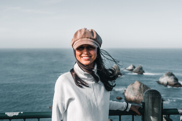 Canvas Print - brunette girl with long hair moved by the wind white sweater corduroy hat looking at camera smiling happily from the viewpoint near the sea, nugget point, new zealand