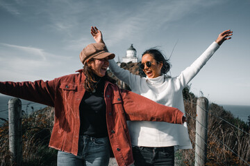 Canvas Print - two caucasian brunette girls come from the lighthouse running happy and smiling with arms outstretched jumping down the dirt road, nugget point, new zealand