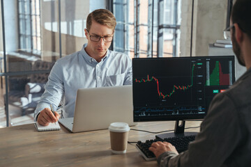 Two stock traders co-working at office sitting at table in front of computers opposite each other, working online, analyzing stock exchange market tendency, weighing risks, selling or buying crypto