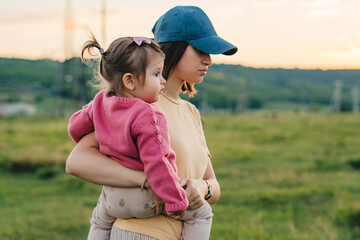 Wall Mural - Side view of a woman holding her little girl in her arms and walking through nature. Activity relationship. Happy family, childhood. Summer nature. Parent