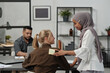 © pressmaster - Young smiling Muslim woman in hijab putting notepaper with word fool on back of blond businesswoman sitting by workplace