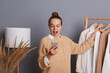 Excited woman shopper wearing beige knitted sweater standing near shelves with clothes choosing new outfit, holding smart phone, looking at display with amazement, female doing shopping.