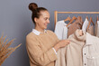 Attractive smiling woman with dark hair and bun hairstyle holding clothes on racks in modern boutique, choosing attire, trying to make stylish trendy look, expressing happiness.