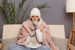 Indoor shot of unhealthy ill woman sitting on the sofa at home wearing winter coat, hat and mittens, cold female suffering flu? coughing, covering mouth with hand.