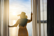 Rear Back View Woman Opening Curtains, Relaxing And Looking Beautiful Seascape Panorama With Rays Of Sun Light In Holiday On The Balcony In High Quality Hotel Feeling Happy. Composition Of Nature