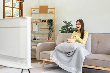 Wall Mural - Young asian woman putting a bowl of popcorn on pillow and eating popcorn while sitting on the big comfortable