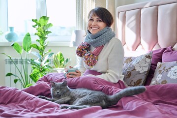 Wall Mural - Woman sitting at home in bed, warming with scarf and mug, with gray cat