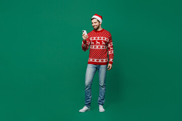 Poster - Full body merry young man 20s in red warm knitted sweater Santa hat posing hold in hand use mobile cell phone isolated on plain dark green background studio New Year 2023 holiday celebration concept