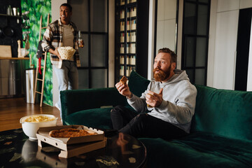 Poster - Two male friends holding beer and pizza while watching sport match in living room