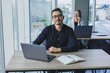 © Дмитрий Ткачук - Attractive business man of Caucasian appearance in a black shirt sits at a desk using a laptop in a modern office with colleagues in the background. Working atmosphere in the office.