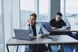 © Дмитрий Ткачук - A female manager is sitting at a desk using a laptop in a modern office against the background of a colleague. Working atmosphere in the office.