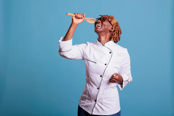 Wall Mural - African american female professional chef wearing kitchen uniform singing excitedly with wooden spoon. Female cook relaxing after work in studio shot against blue background.