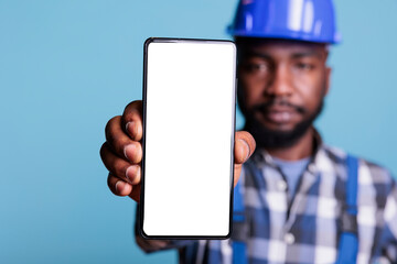 Confident african american contractor holding cell phone with white screen mockup design wearing coveralls in studio. Builder using touchscreen device for advertising construction industry concept.