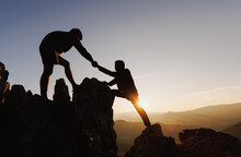 Silhouette Of  Two People Climbing On Mountain Cliff And One Of Them Giving Helping Hand. People Helping And, Team Work Concept.