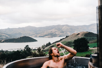 Wall Mural - caucasian young man inside wooden hot tub with outstretched hand and open mouth in the middle of the forest near the peaceful trees and greenery in full nature, te wepu pods akaroa, new zealand