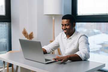 Poster - Cheerful young African-American male entrepreneur in casual clothes working on business project on laptop sitting at desk in modern office room with light interior, on background of window.