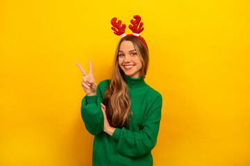 Happy smiling brunette girl showing peace gesture or v-sign with two fingers, wearing knitted green sweater with red Christmas reindeer antlers and looking at camera on a yellow background.