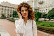 © PhotoBook - Close-up photo of pleasant young curly caucasian lady spends weekend outdoors. She touches her hair and looking at camera against old city building. Lifestyle concept