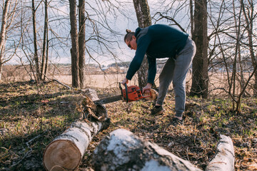 Preparation for the heating season, firewood harvesting. A young man with a chainsaw sawing a fallen tree in the forest.