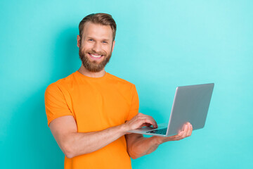 Portrait of handsome positive glad guy with blond hairdo wear orange t-shirt hold laptop write email isolated on teal color background