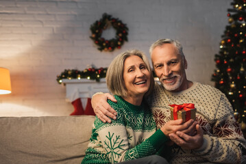 Wall Mural - happy middle aged man in sweater hugging cheerful wife with christmas present