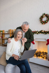 Wall Mural - cheerful middle aged woman in glasses using laptop and holding hands with happy husband during christmas holidays