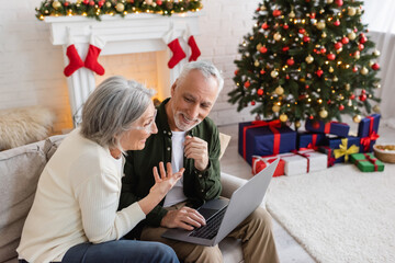 Wall Mural - smiling middle aged woman pointing at happy husband and having video call on laptop during christmas