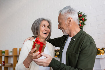 Wall Mural - smiling mature woman holding christmas present near cheerful husband