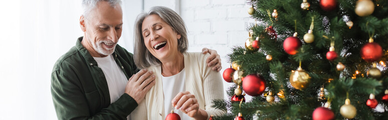 Wall Mural - cheerful middle aged woman laughing while holding bauble near husband and christmas tree, banner