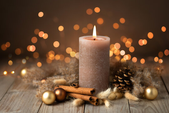 Fototapete - Warm colored natural Christmas Advent candle still life. Brown candle decorated with cinnamon sticks, pine cone, golden balls and dried grass in Boho style on rustic wood with Bokeh lights.