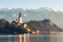 Beautiful Shot Of The Lake Bled With A Church And A Castle In The Background With Snowy Mountains