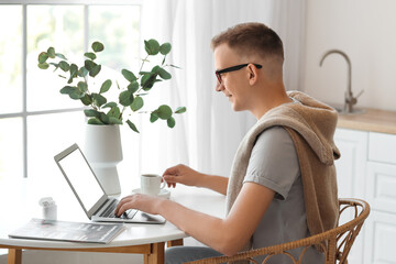 Poster - Young man with cup of coffee using laptop at table in office