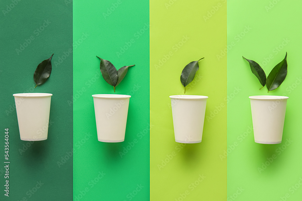 Paper cups with plant leaves on color background