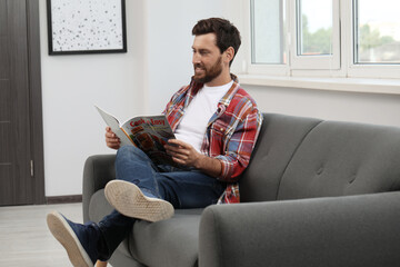 Canvas Print - Smiling bearded man reading magazine on sofa at home