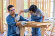 © Bangkok Click Studio - Asian male carpenter woodworker engineer dad in jeans outfit with safety gloves and glasses goggles helping teaching son using pencil marking wood stick board plank on workbench in construction site