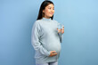 Pregnant woman wearing winter sweater drinking glass of water isolated on blue