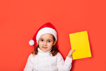 Wall Mural - Cute caucasian kid girl in red Santa's hat holding certificate or greeting card empty copy space for design or text. Banner. Christmas New Year mockup template.