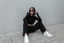Fashionable Young Urban Woman With Black Trendy Sunglasses In Fashion Black Stylish Sports Outfit With White Sneakers Sits On The Street Near A Modern Gray Building