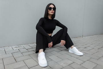 Beautiful woman with bob hairstyle with black fashion sunglasses in black sports stylish outfit with white sneakers sits near a gray building on the street