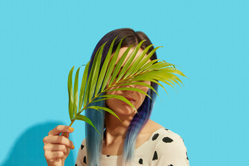 Wall Mural - Young beautiful woman with blue hair holding palm leaf on bright background