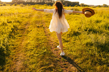 Wall Mural - Rear view of unrecognizable cheerful woman in straw hat and white dress joyfully running bouncing along in rural road field toward sun in summer day. Happy female enjoying freedom on sunny meadow.