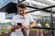 © Miljan Živković - One caucasian man taking a brake during outdoor training in the park outdoor gym using mobile phone texting or reading typing resting on the bars with supplement shaker in hand happy smile copy space