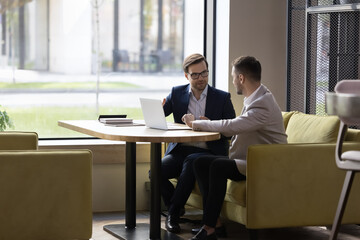 in cozy office two millennial businessmen in suits having formal talk over collaborative on-line pro