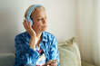 © evafesenuk - Senior woman in denim clothes in blue headphones sits on the couch at home listening to music