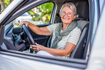 Sticker - Portrait of attractive happy senior woman in t-shirt sitting in her car, with hands holds steering wheel, looking at camera smiling