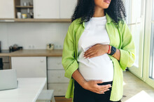No Face Picture Of Pregnant African American Woman With Long Curly Hair Looking Through Window Standing In Kitchen, Rubbing Her Big Cute Belly, Expecting Baby Boy To Be Born In Few Month