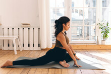 Prenatal fitness training. Sporty african american pregnant woman in leggings sitting on left leg, stretching right, practicing prenatal yoga or pilates on mat at home against window