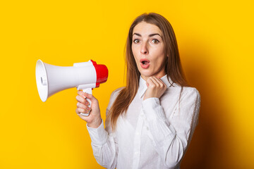 Surprised young woman in a white shirt holds a megaphone on a yellow background. Hiring concept, ad